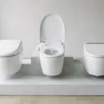 Three white bidet toilet seats displayed side by side on a tiered platform in a bright bathroom showroom